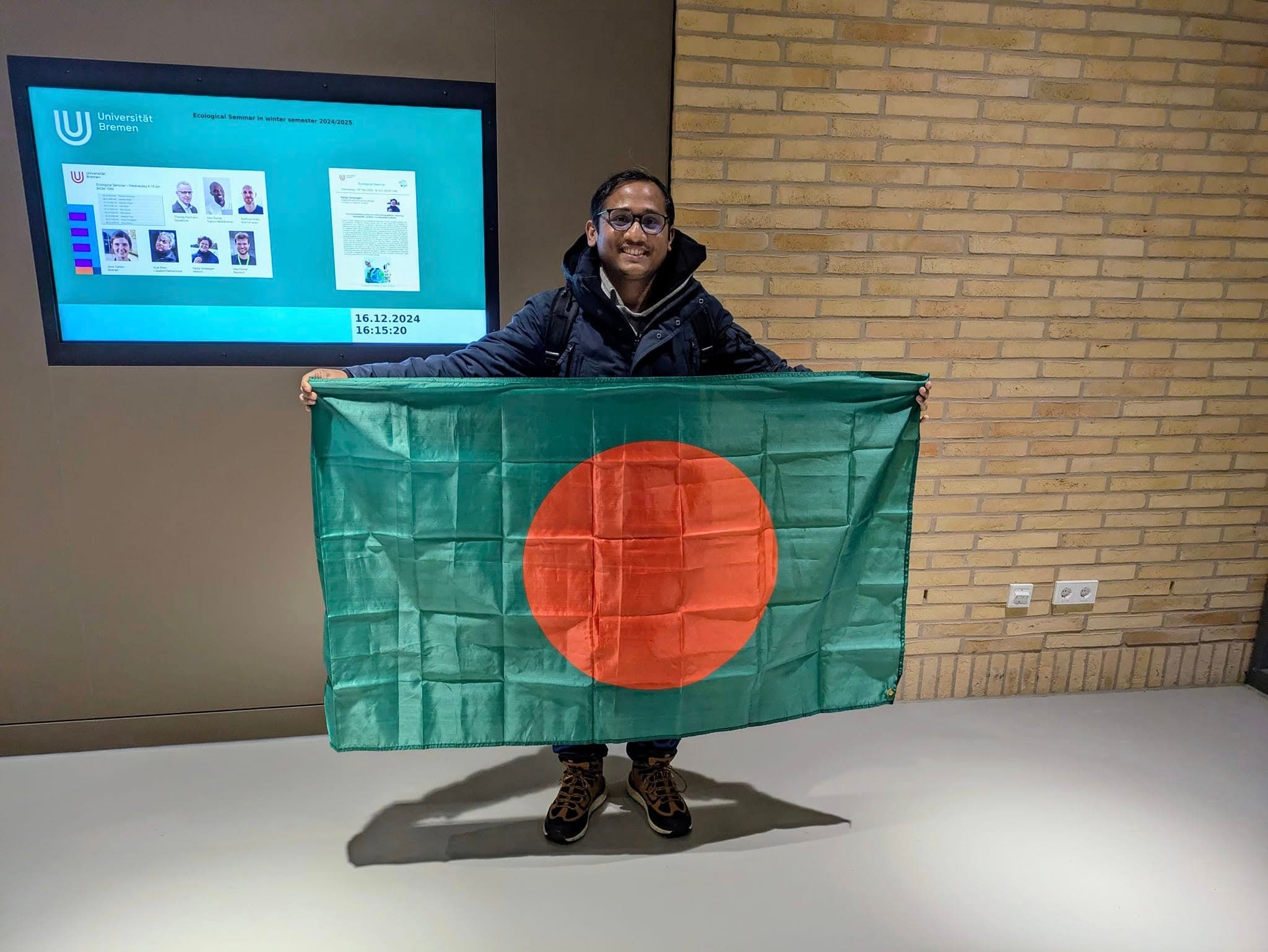 Uachash Barua at the University of Bremen holding the flag of Bangladesh beside an ecological seminar display